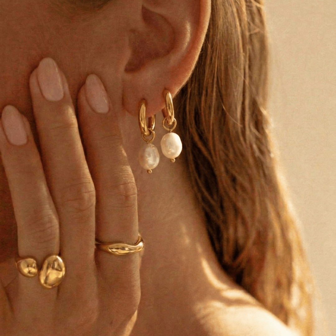 Pendientes de argolla dorados con perla blanca; mano y oreja en primer plano
